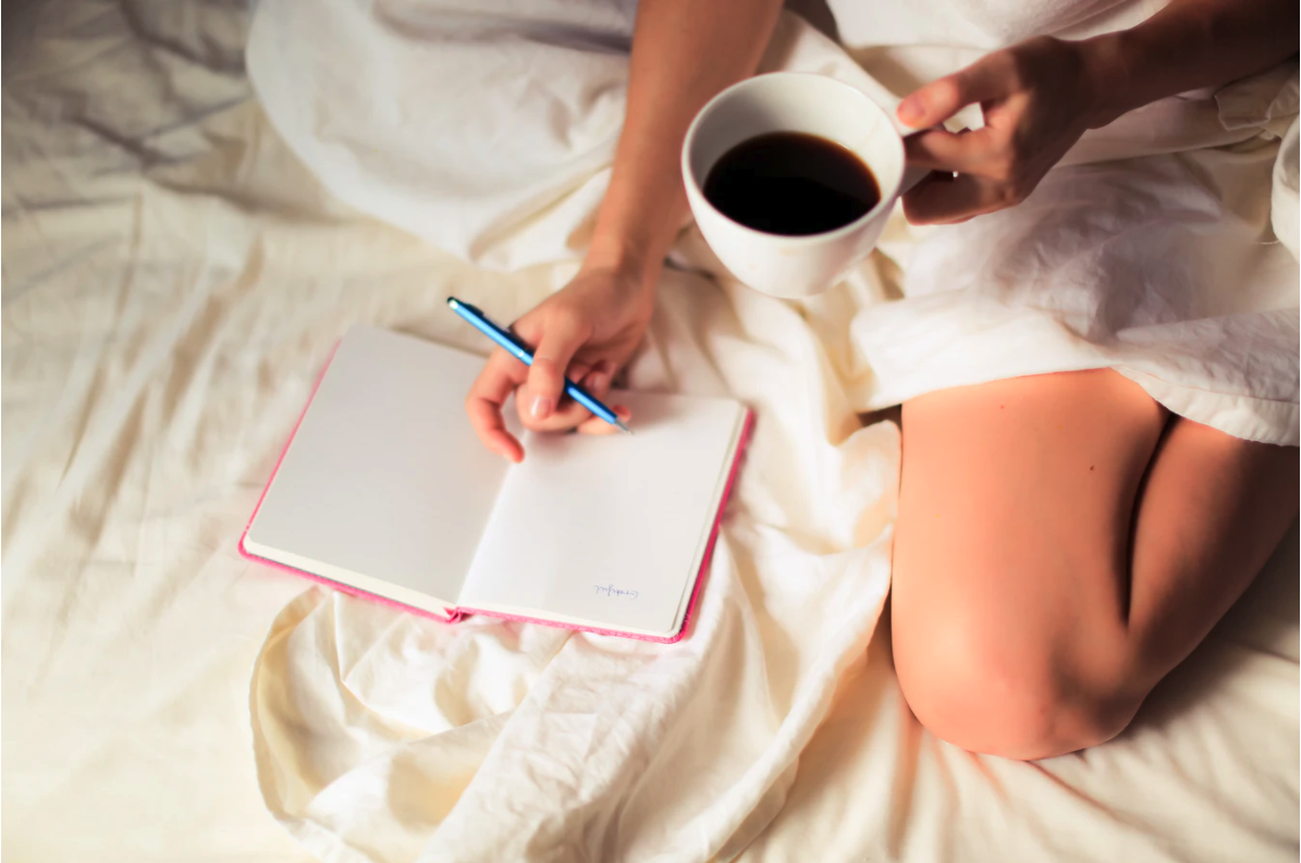 A woman in bed with a cup of coffee writing in a notebook. An example of staying positive while at home.